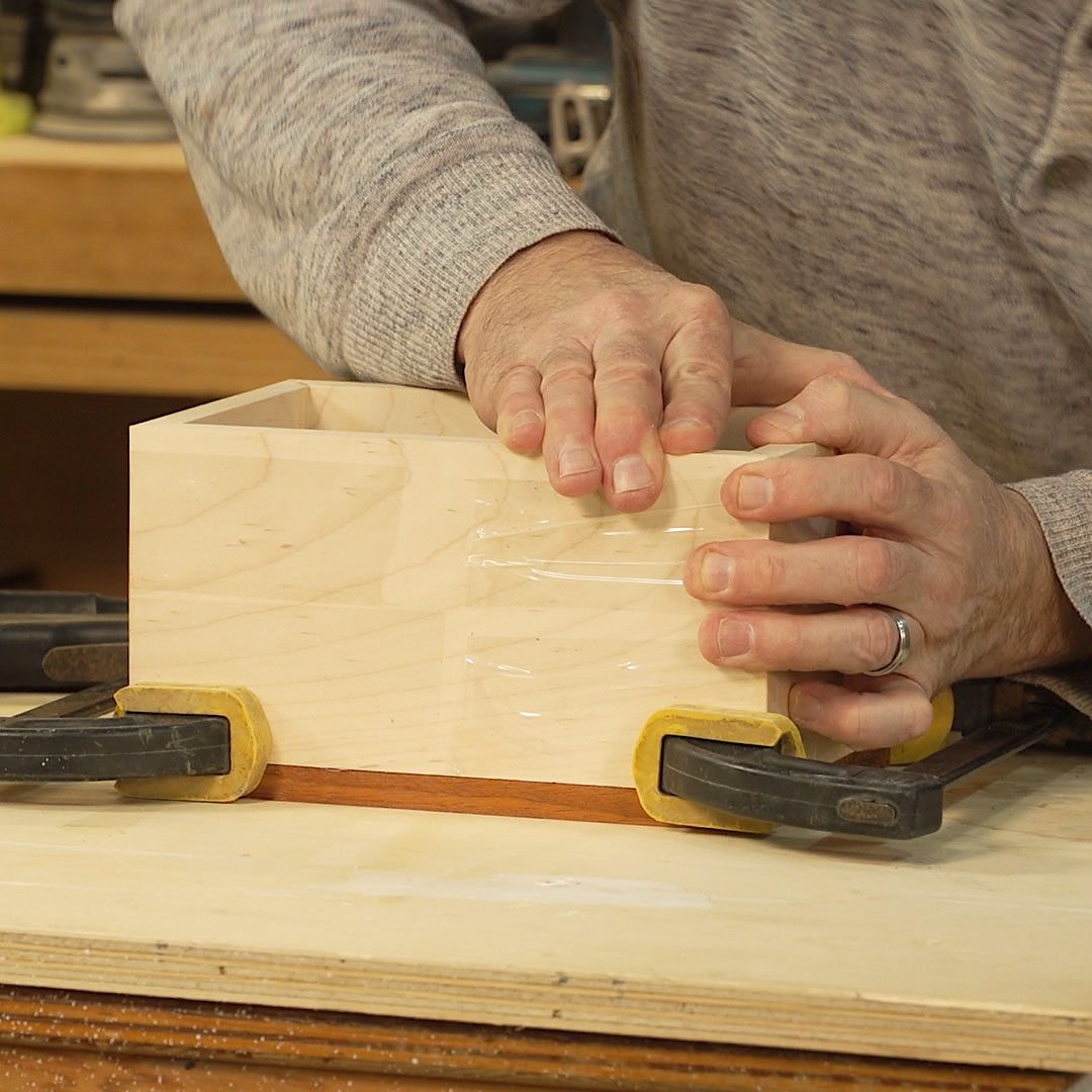 A woodworker clamps a rectangular wooden box together, pressing down firmly with both hands, on a workbench surrounded by tools and materials.