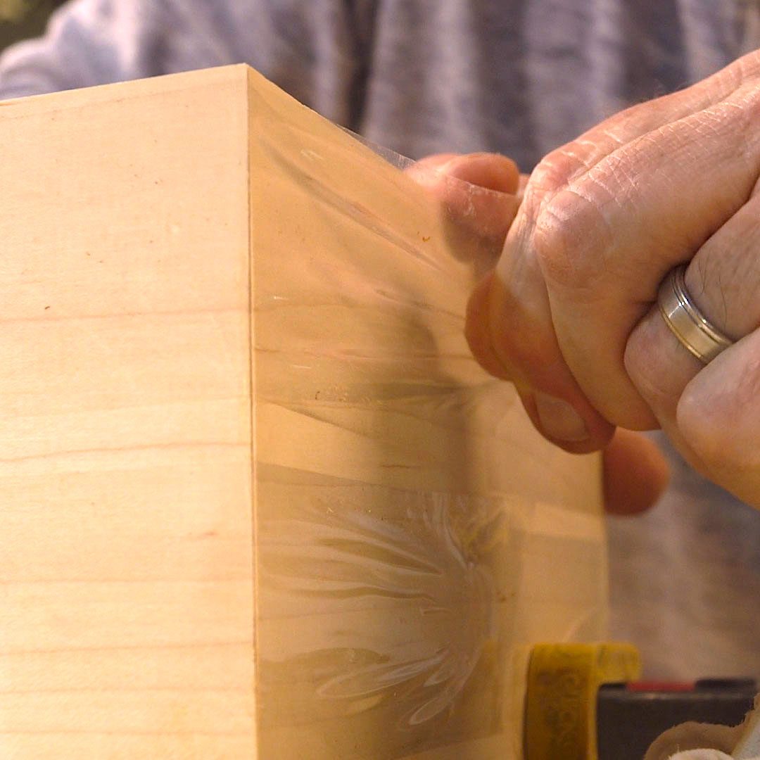 A hand smooths a glossy surface of a wooden block, surrounded by tools like a tape measure, in a workshop setting.