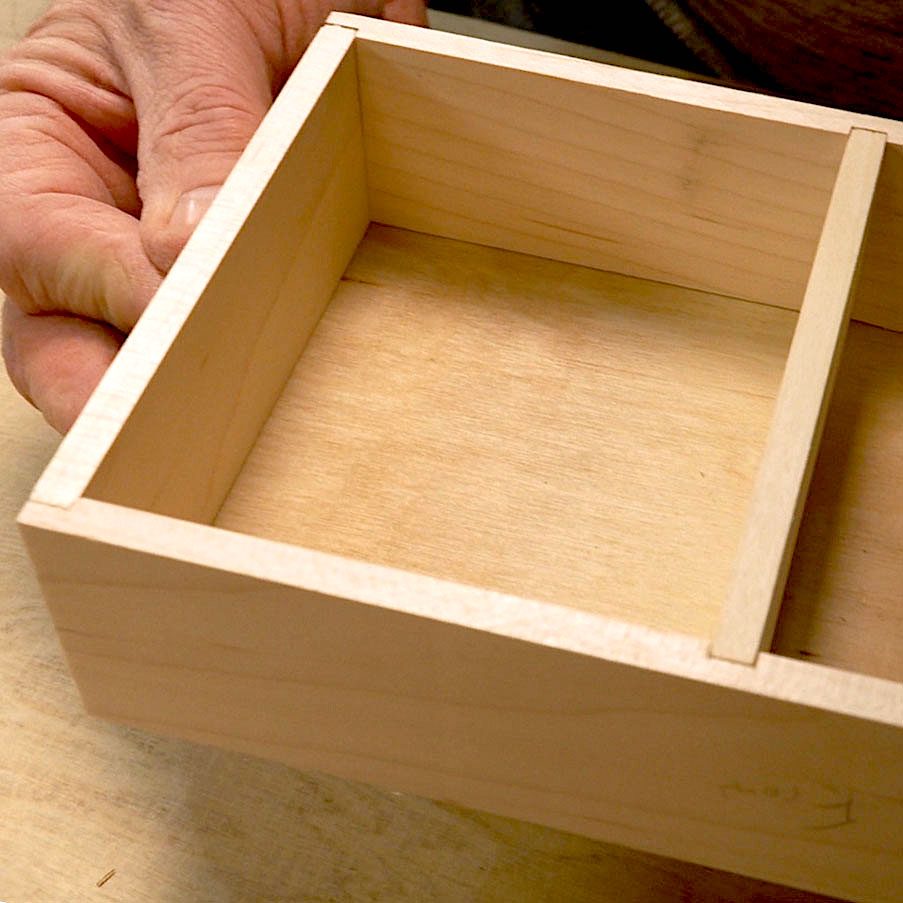 A hand holds a wooden box with an interior divider, suggesting preparation for organization or storage on a work surface.