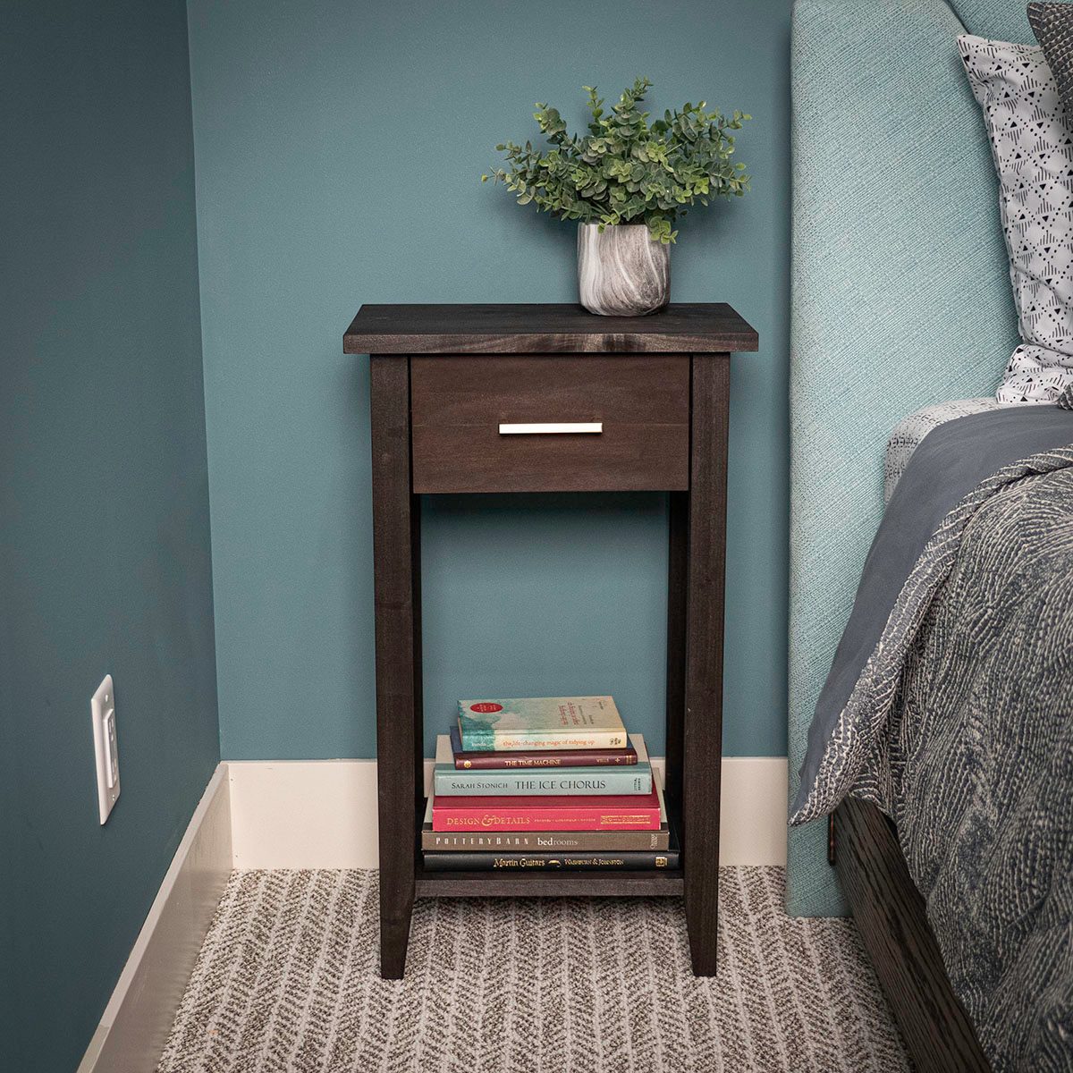 A dark wood nightstand stands beside a bed, featuring a drawer, stacked books underneath, and a small plant atop against a teal wall.