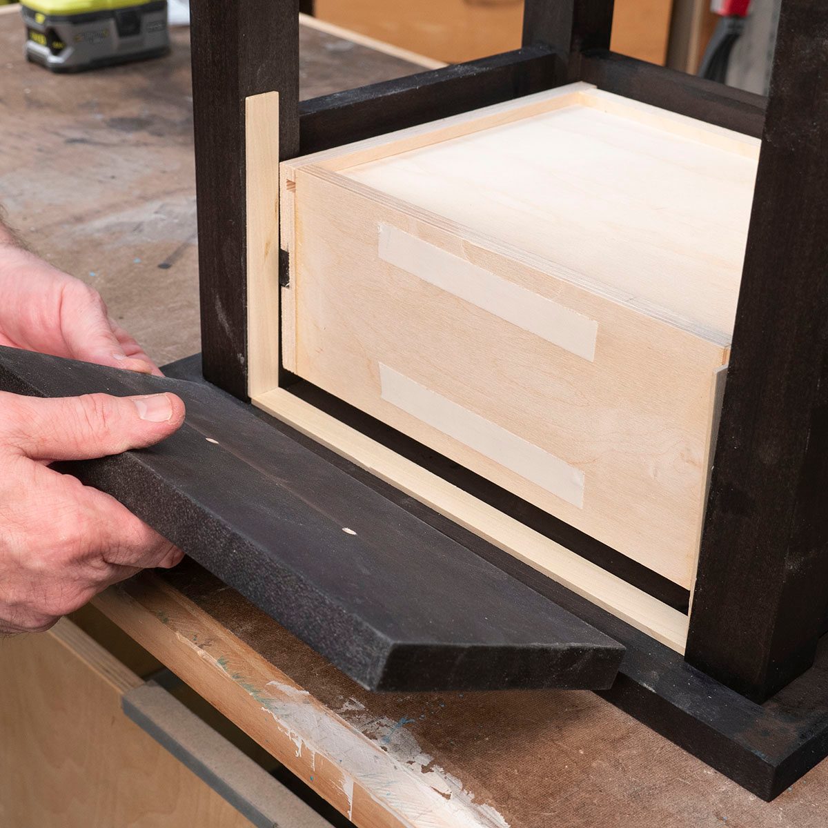 A person holds a dark wooden piece, aligning it with a drawer in a wooden frame, situated on a workbench with various tools in the background.