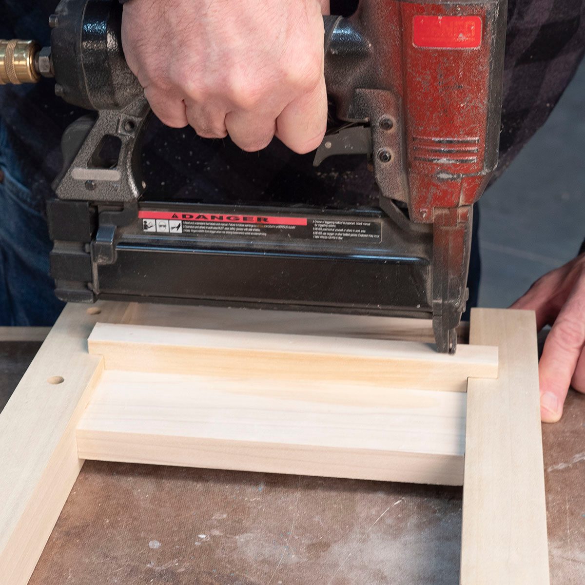 A hand holds a nail gun, driving nails into wooden pieces on a workbench, contributing to an assembly project in a workshop environment.