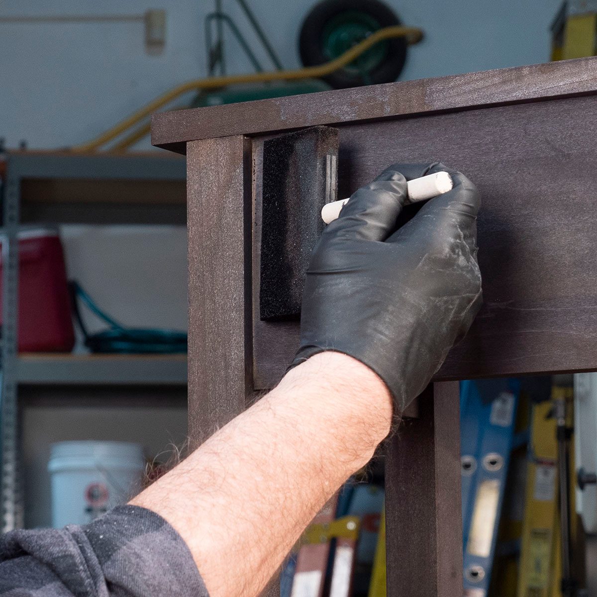 A hand wearing a black glove writes with a white marker on a dark wooden surface, surrounded by tools and storage items in a workshop.