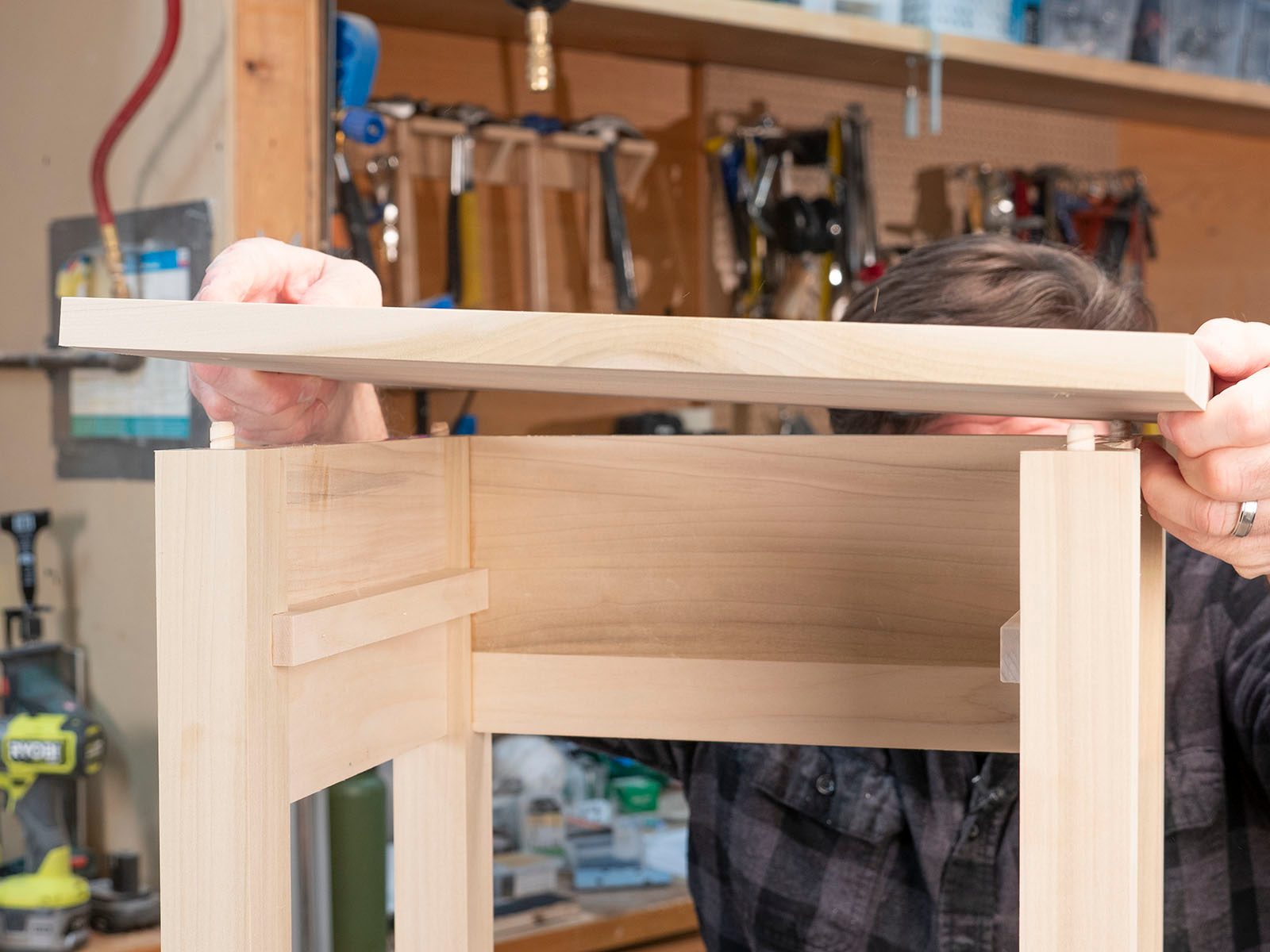 A person is placing a wooden tabletop onto a partially assembled table, surrounded by various woodworking tools in a workshop environment.