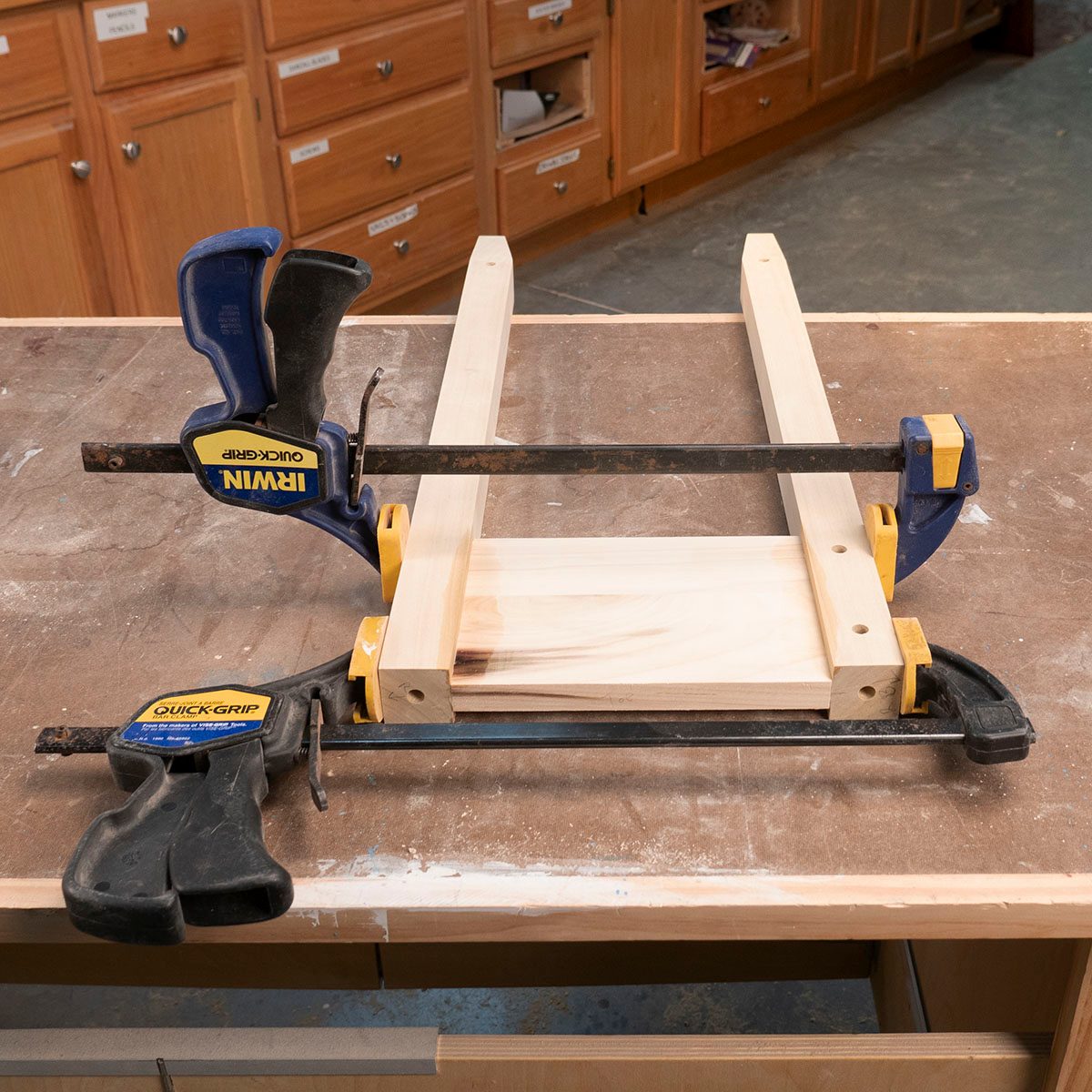 Two clamps secure wooden pieces on a workbench, preparing them for assembly. The background features wooden cabinets with labeled drawers.