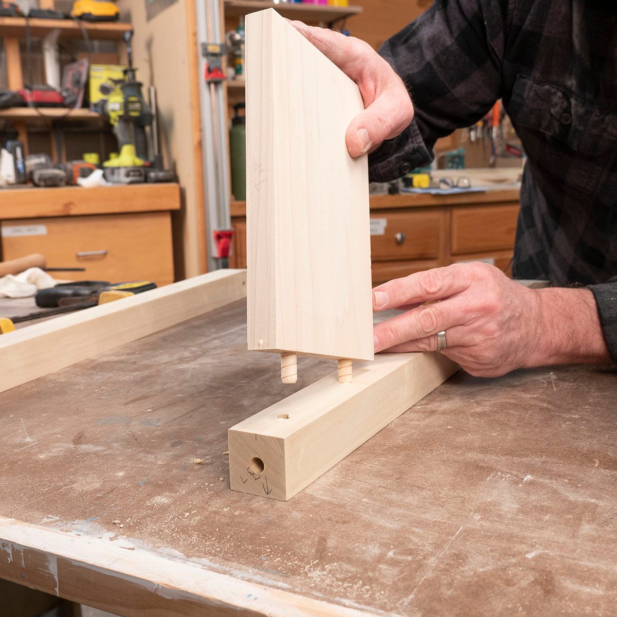 A person aligns a wooden panel with pegs into a corresponding slot on a larger wooden piece, surrounded by tools and materials on a workbench.