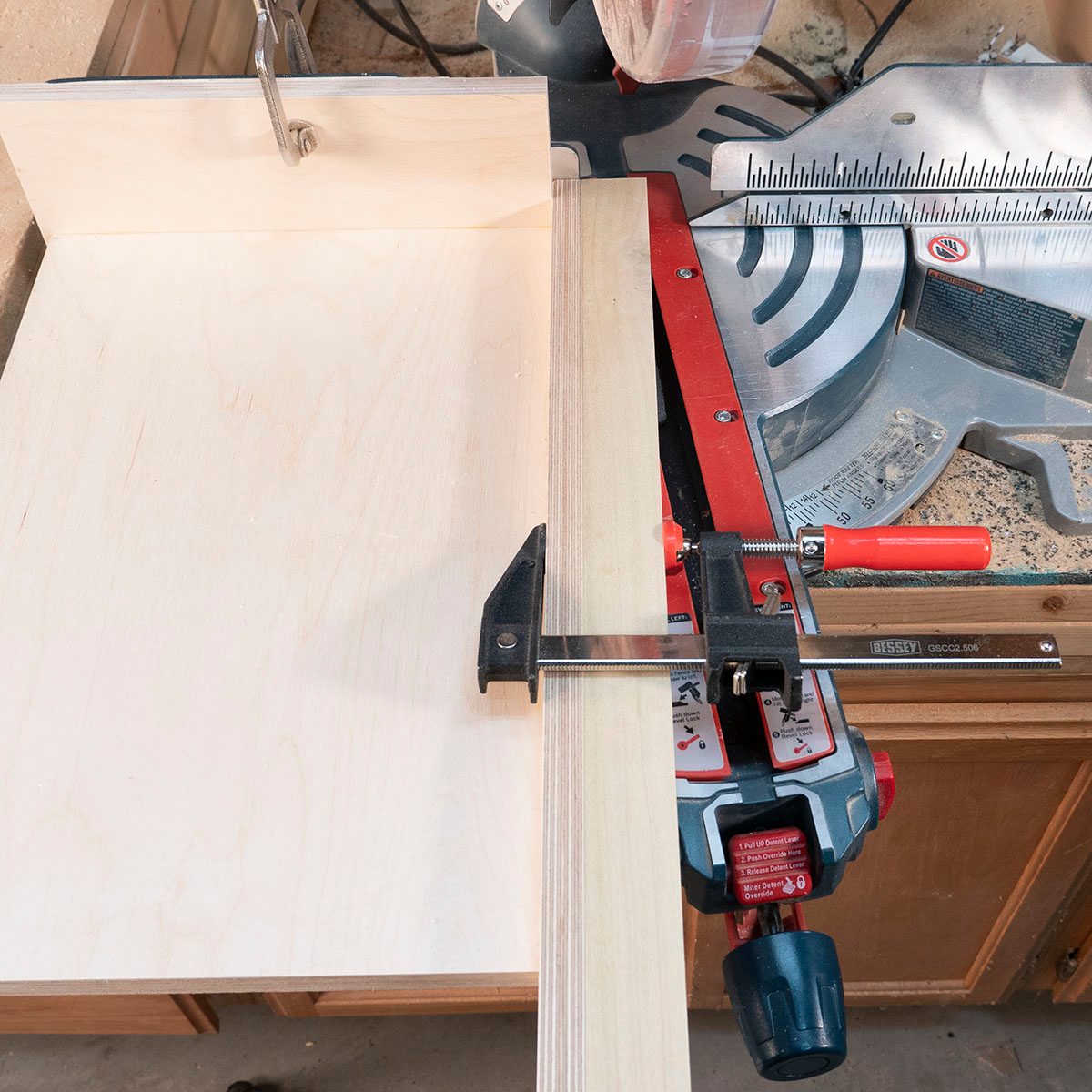A wooden panel is clamped against a miter saw's fence, positioned for cutting in a workshop with visible tools and surfaces.