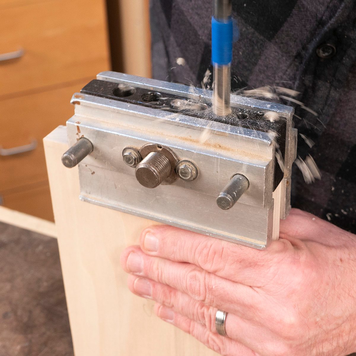 A hand holds a metal drilling guide against wood while a drill bit rotates, creating shavings and dust in a woodworking environment.