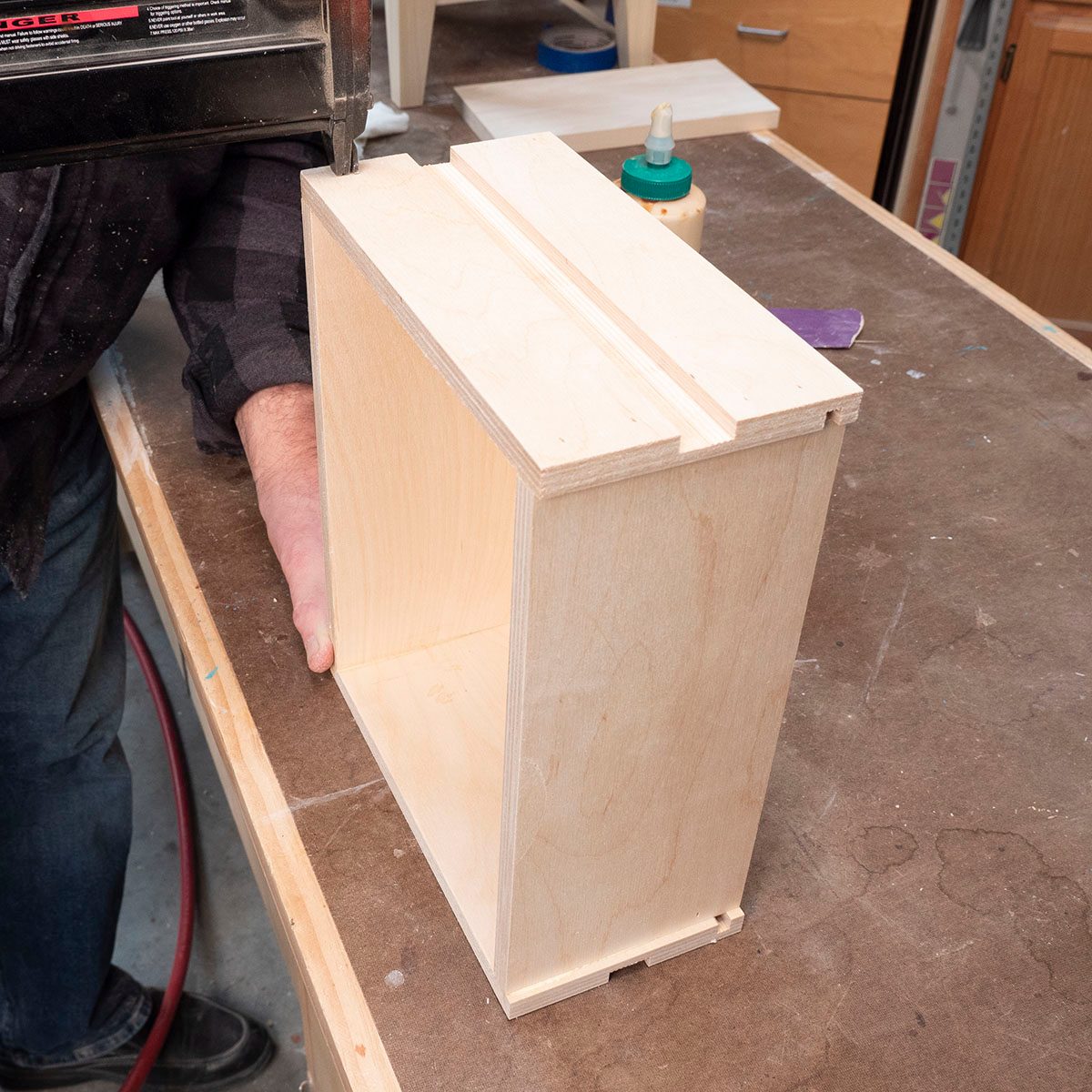 A person holds a wooden box, using a nail gun to secure one end. The setting is a workshop with tools and wood surfaces around.
