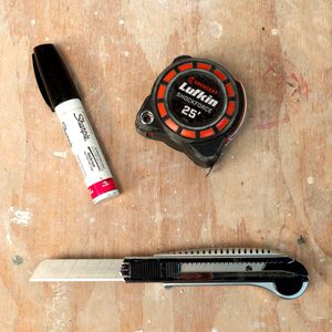 A tape measure, marker, and utility knife rest on a wooden surface, suggesting preparation for a measurement or cutting task in a workshop setting.