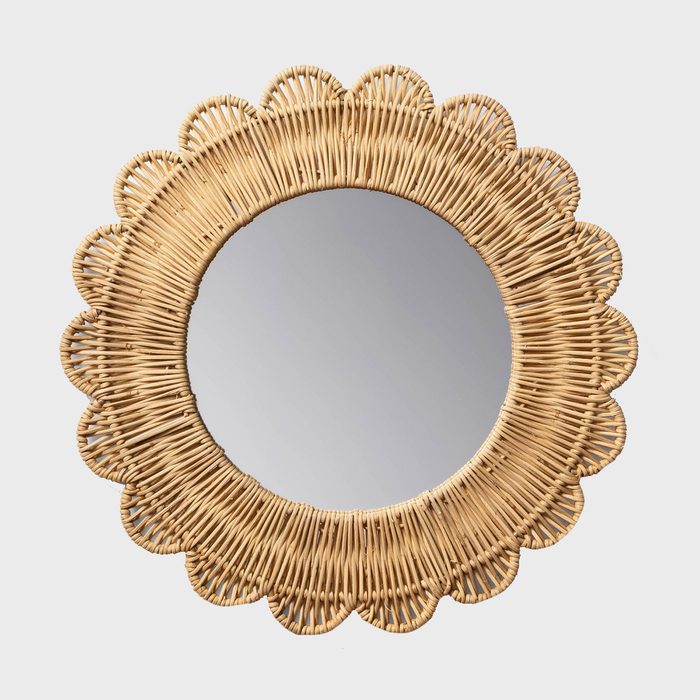 A round mirror is surrounded by a textured, woven rattan frame with a scalloped edge, reflecting light in a clean, neutral background.