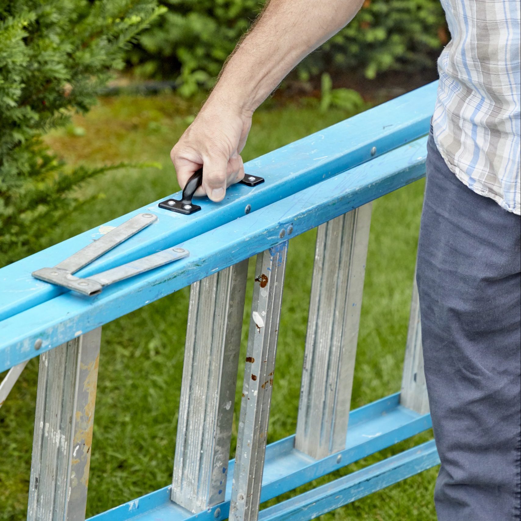 A hand grips a black handle on a blue ladder, adjusting it on green grass surrounded by shrubbery, suggesting preparation for a task.