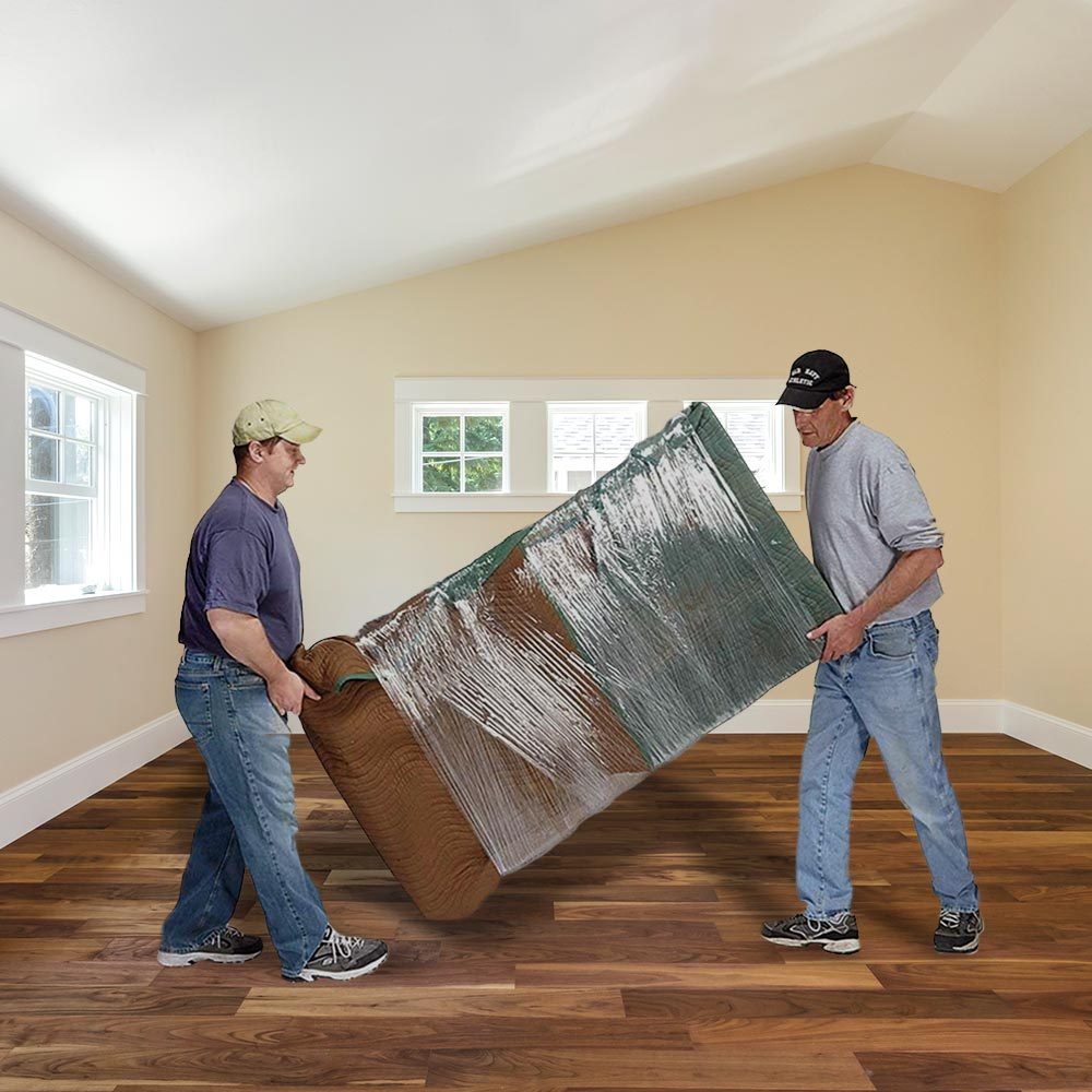 Two men lift a large, wrapped object, likely a piece of furniture, in a bright, empty room with wood flooring and windows.