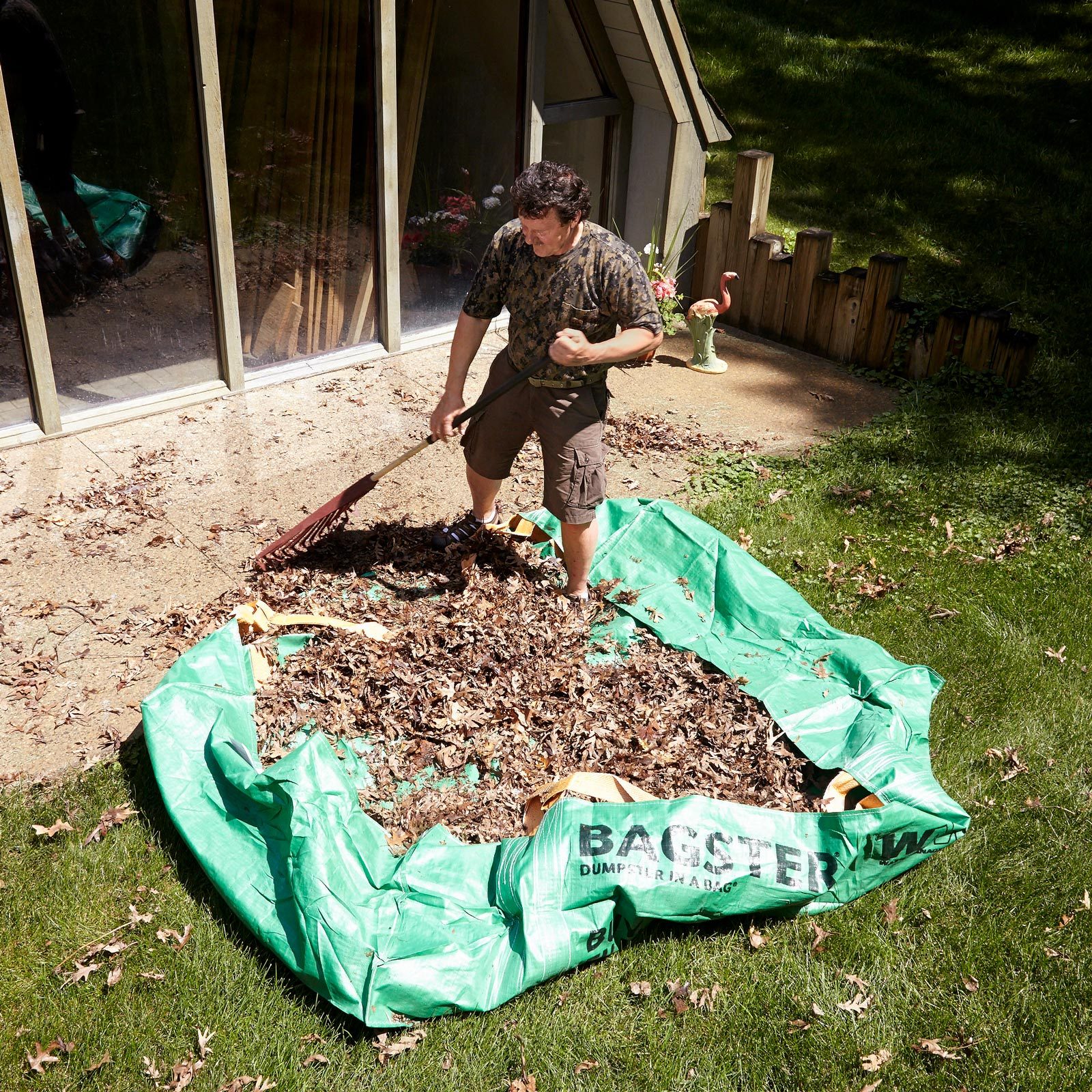 A man rakes leaves into a large green bag in a sunny backyard, near a house with large windows and decorative items.