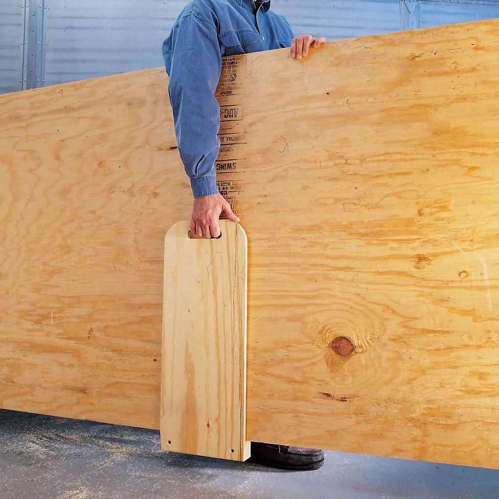 A person lifts a large wooden plank using a handhold, standing on a wooden floor with sawdust in a workshop setting.
