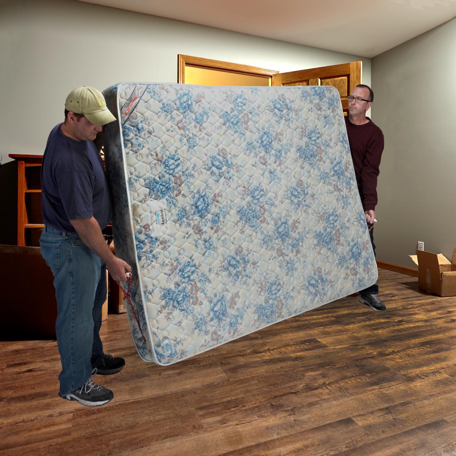 Two men are lifting a large floral-patterned mattress in a room with wooden flooring, a doorway, and furniture in the background.
