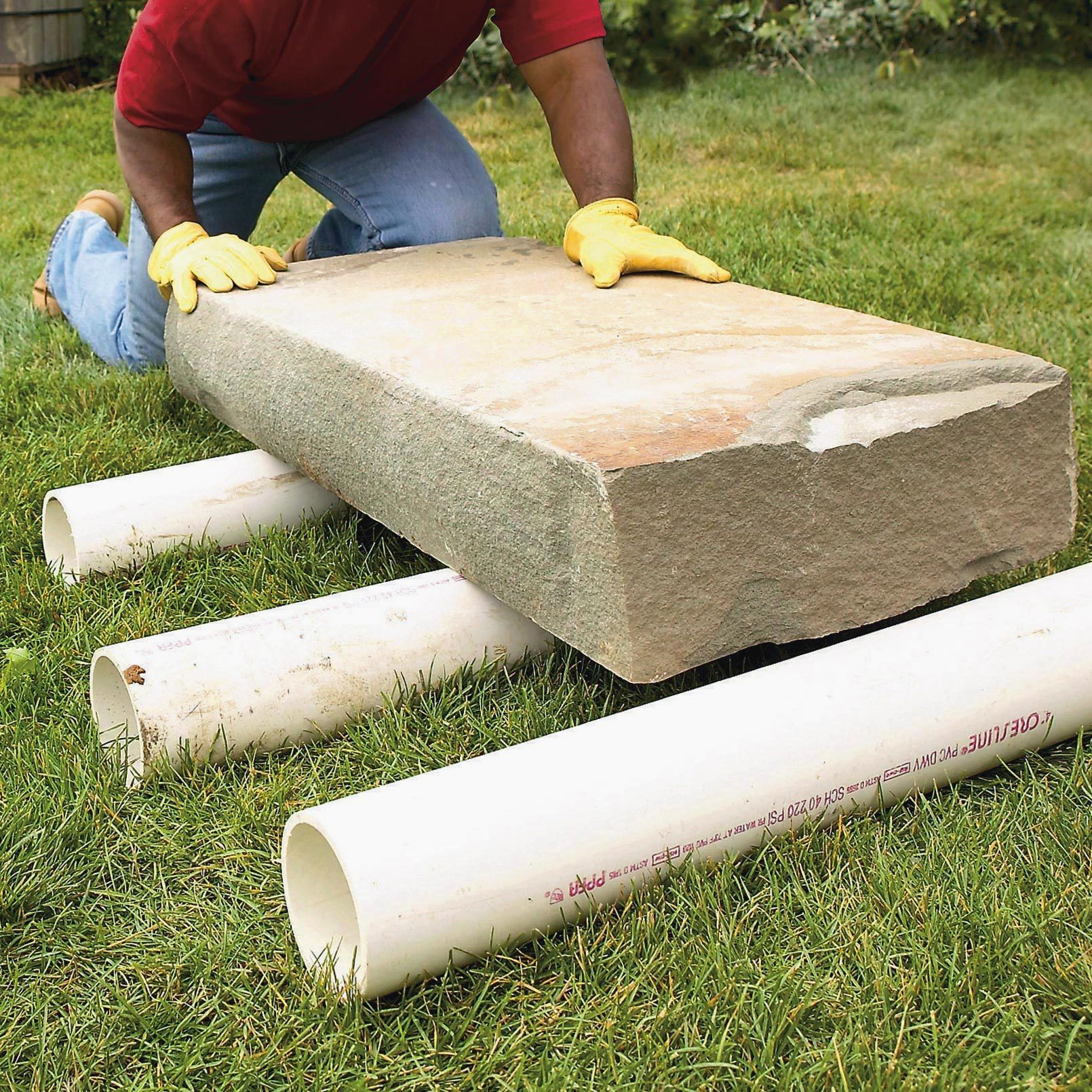 A person places a large stone onto three white PVC pipes on grassy ground, using gloves to assist in positioning the heavy object.