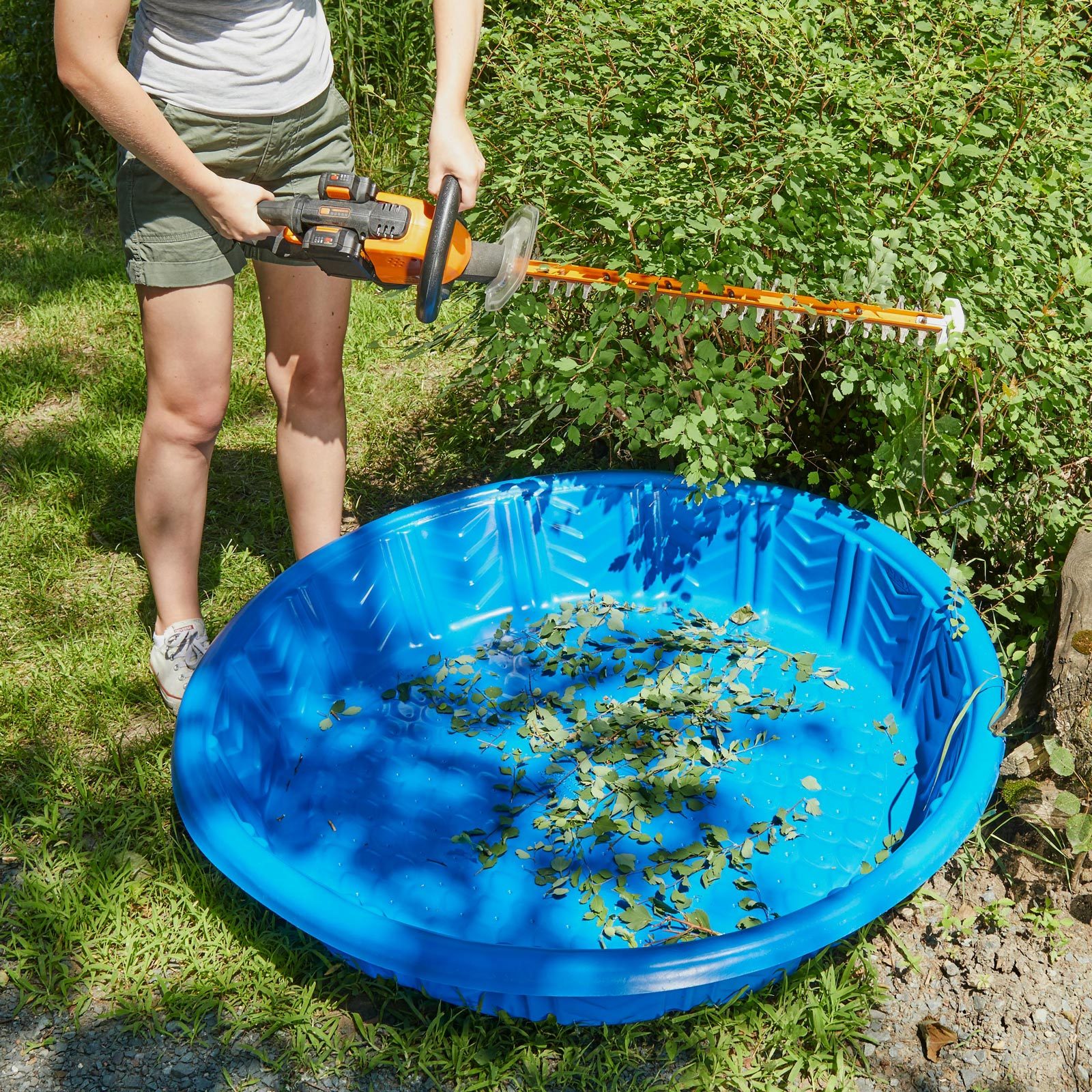 A person trims bushes with a hedge trimmer above a blue plastic pool containing leaves, set in a grassy area surrounded by greenery.
