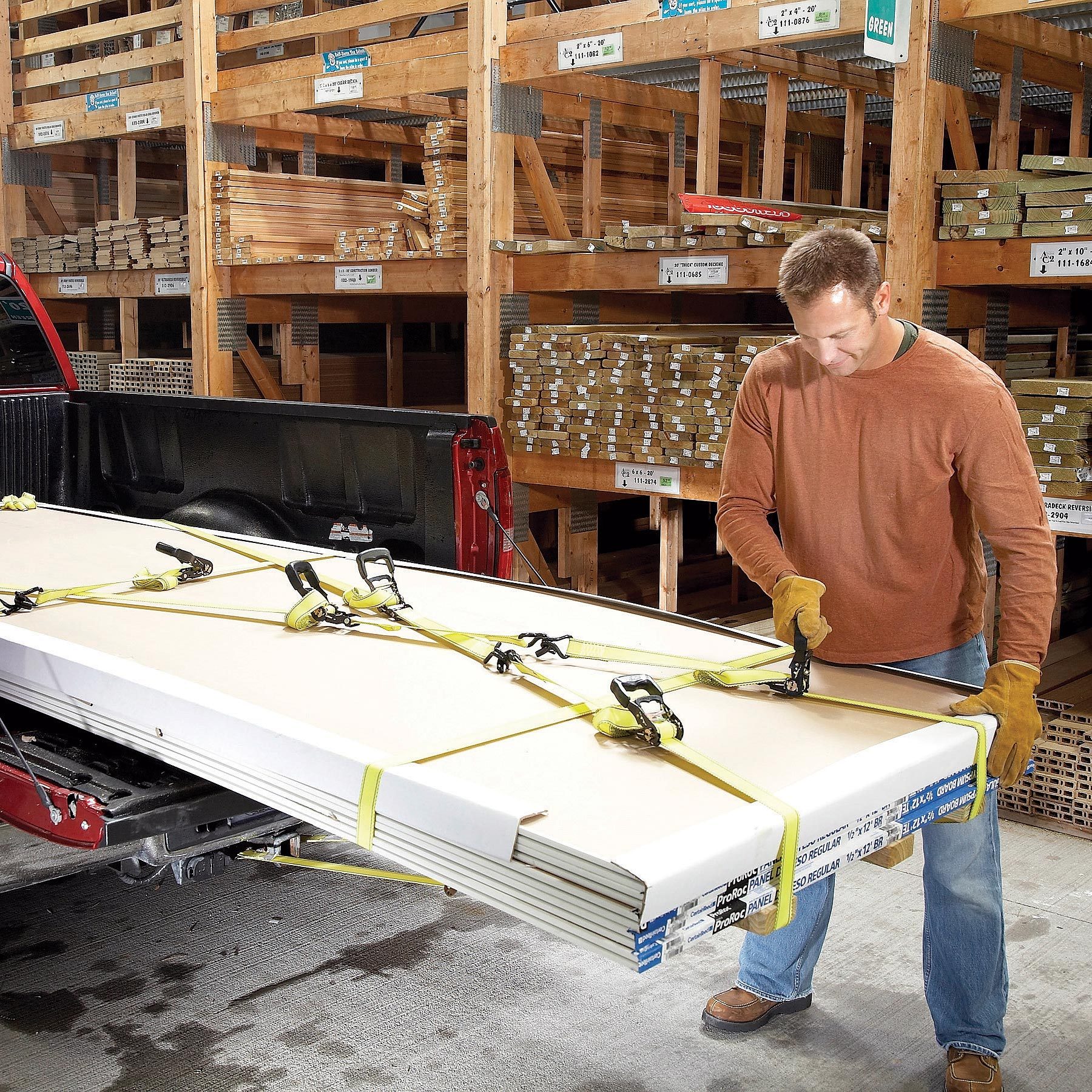 A man secures large sheets of material in a truck bed using straps and clamps inside a warehouse filled with stacked wooden materials.