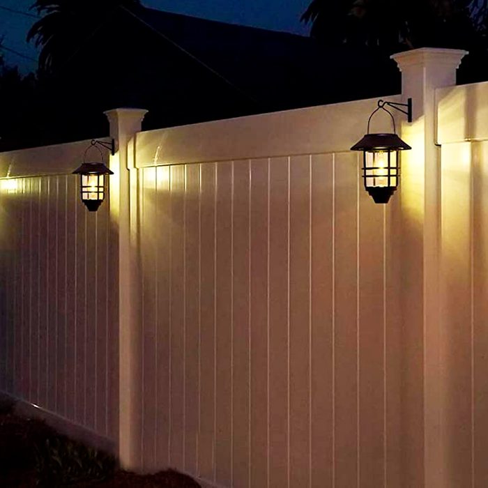 A white wooden fence stands illuminated by hanging lanterns, providing light in a dim evening setting surrounded by shadows from nearby structures and trees.