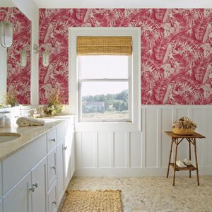 A white bathroom features a marble sink with towels, a wicker basket table, and vibrant pink leaf-patterned wallpaper, illuminated by natural light from a window.