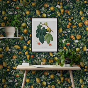 A wooden table displays a book and stone. A framed botanical print hangs on a floral wallpaper, surrounded by green plants on a shelf below.