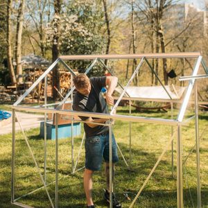 A man is assembling a metal greenhouse frame in a grassy outdoor area, surrounded by trees, a hammock, and various garden items in the background.