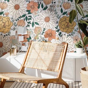 A woven chair sits in sunlight, facing a vibrant floral wallpaper, accompanied by a potted plant and decorative objects on a white cabinet.