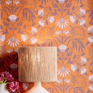 A textured lamp stands on a surface, illuminating against a vibrant orange floral wallpaper featuring intricate flowers and leaves.