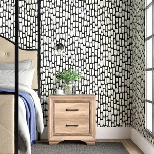 A wooden nightstand with two drawers holds a plant and a mug next to a bed, all set against a patterned black and white wall.