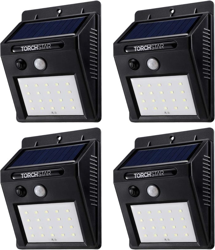 Four solar-powered motion sensor lights are mounted on a wall, designed to illuminate outdoor areas when detecting movement. They have sleek black casings.