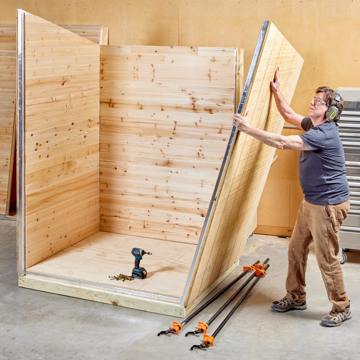 A person is lifting a wooden panel to assemble a wooden structure in a workshop, surrounded by tools and clamps on a concrete floor.