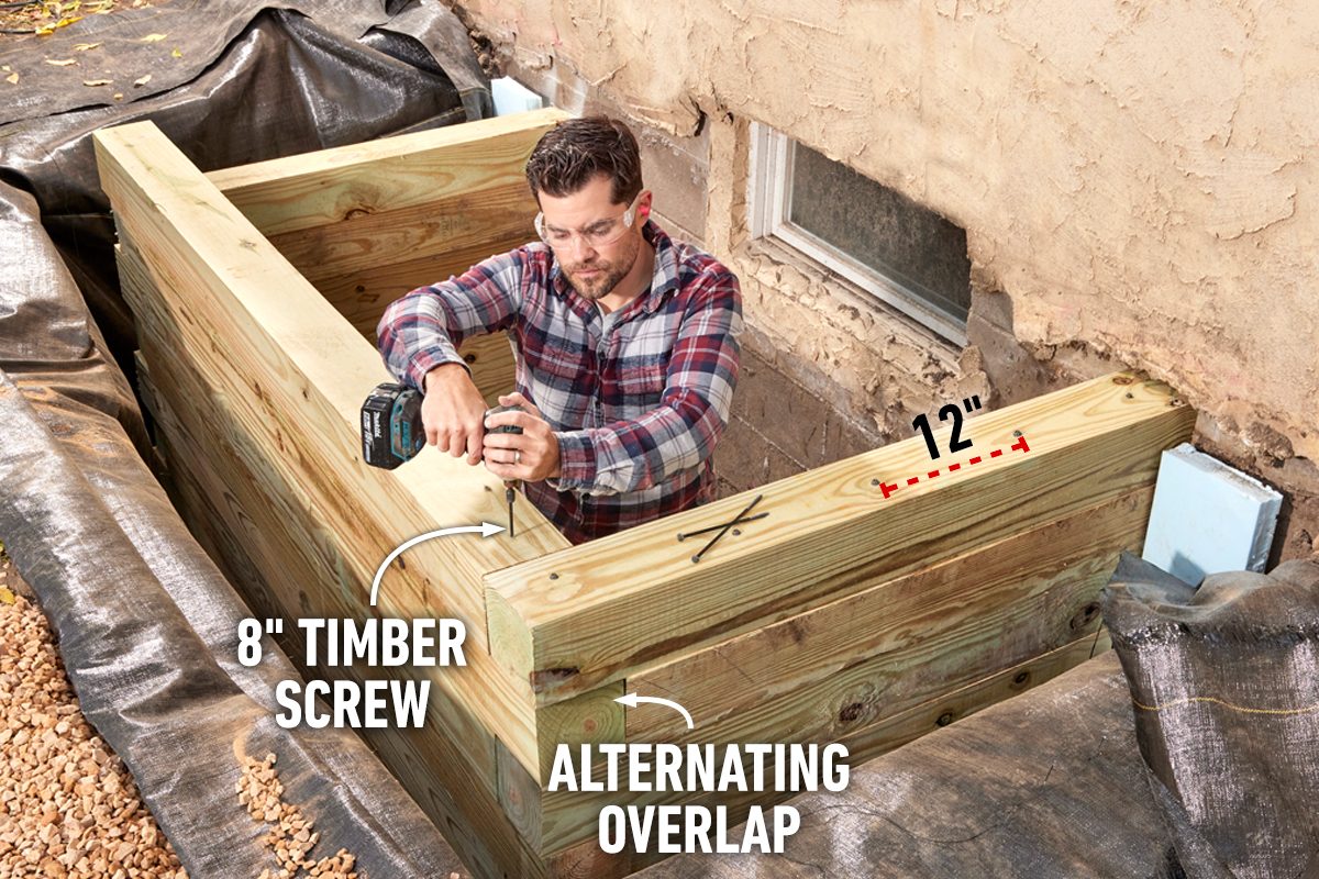 A man uses a drill to secure wooden timbers together in a backyard surrounded by gravel and plastic sheeting against a textured wall.