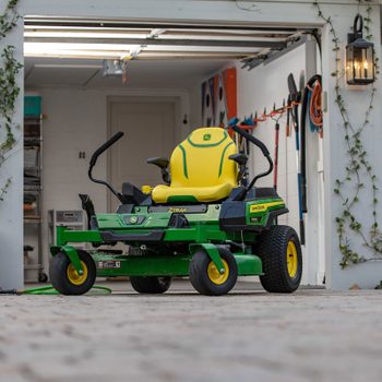 A green and yellow riding lawn mower sits in a garage, surrounded by gardening tools and shelves, with ivy clinging to the walls.