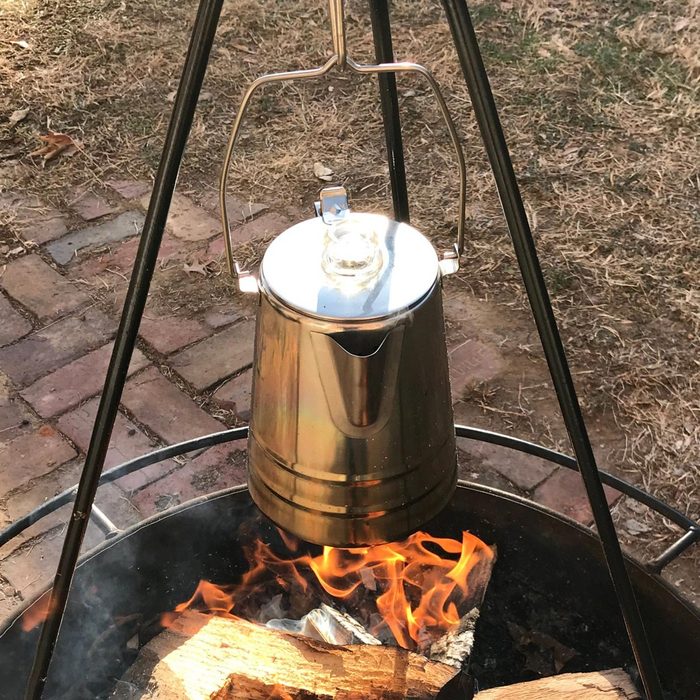 A metal kettle hangs from a tripod over a fire, boiling water amidst dancing flames and glowing logs on a grassy ground.
