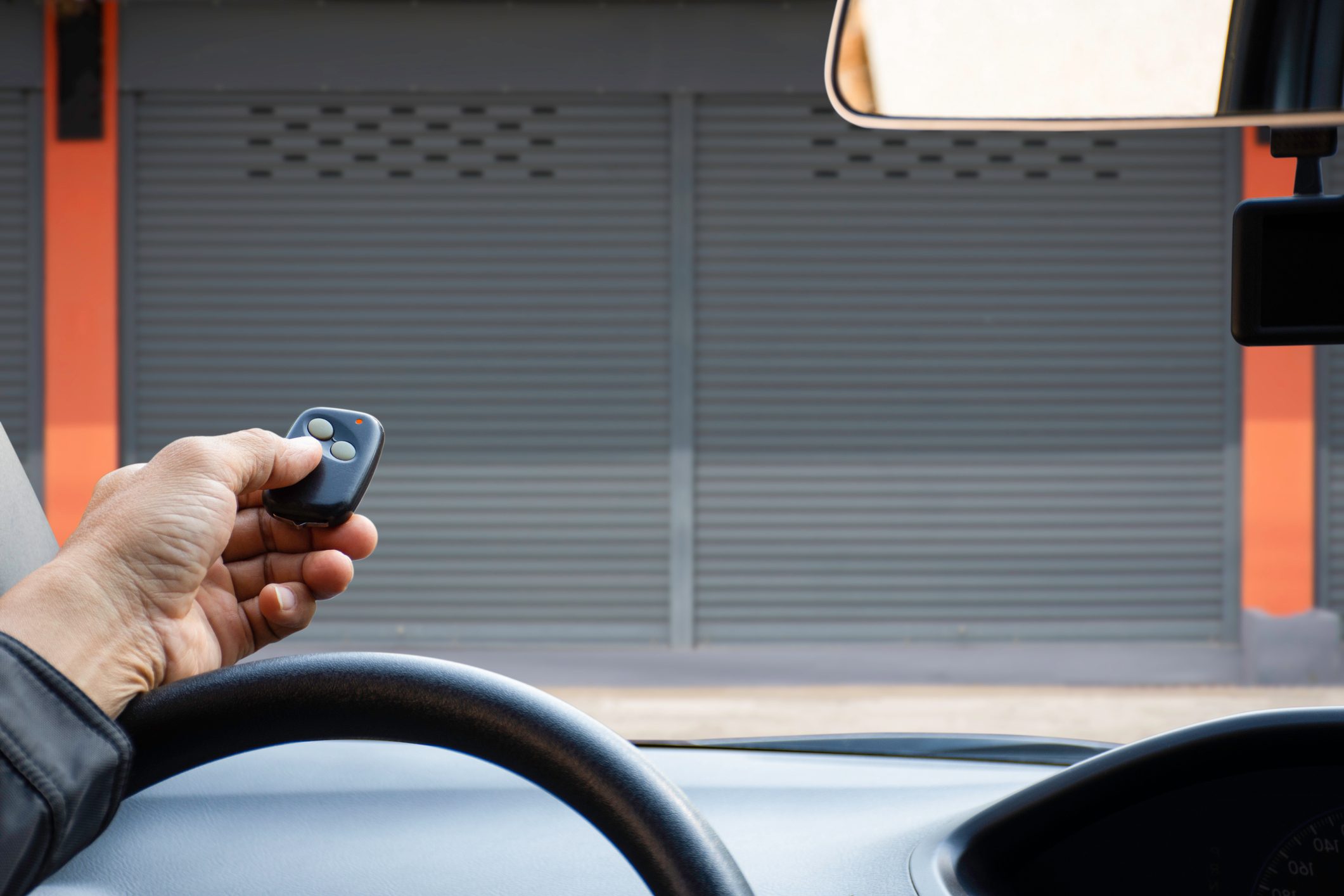A hand holds a remote control, pressing a button inside a car. A closed garage door looms in the background, framed by an orange wall.