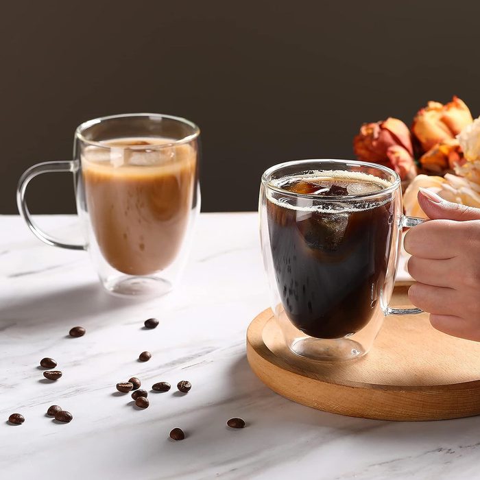Two glass mugs are presented on a countertop; one holds iced coffee while the other contains a creamy beverage, and coffee beans scatter nearby.