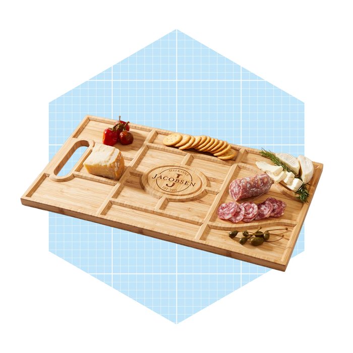 A bamboo cheese board displays assorted cheeses, meats, crackers, olives, and cherry tomatoes, arranged neatly for serving, set against a light blue background.
