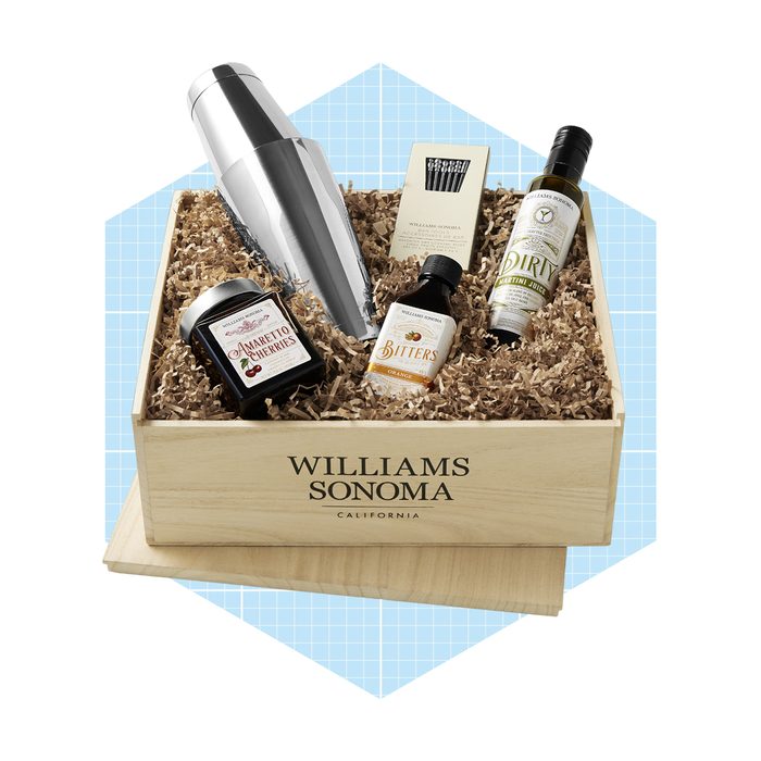 A wooden gift box contains a cocktail shaker, bitters, maraschino cherries, and a martini mix, all arranged in shredded paper, with a light blue background.