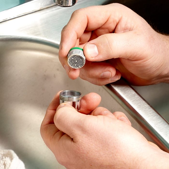 A person holding a disassembled faucet aerator next to a kitchen sink, showcasing the individual components, including the main body and the mesh filter.
