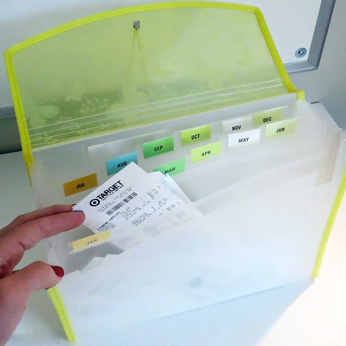 A person holds a receipt while organizing it into a translucent accordion folder with labeled dividers for each month. The outer edge of the folder is yellow, and the background is a white surface.